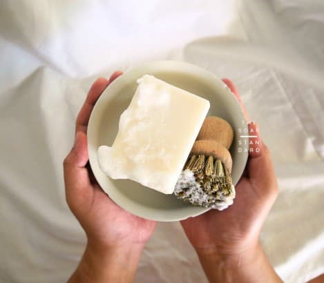 Hands holding a bowl with a sudsy white soap bar and wooden brush